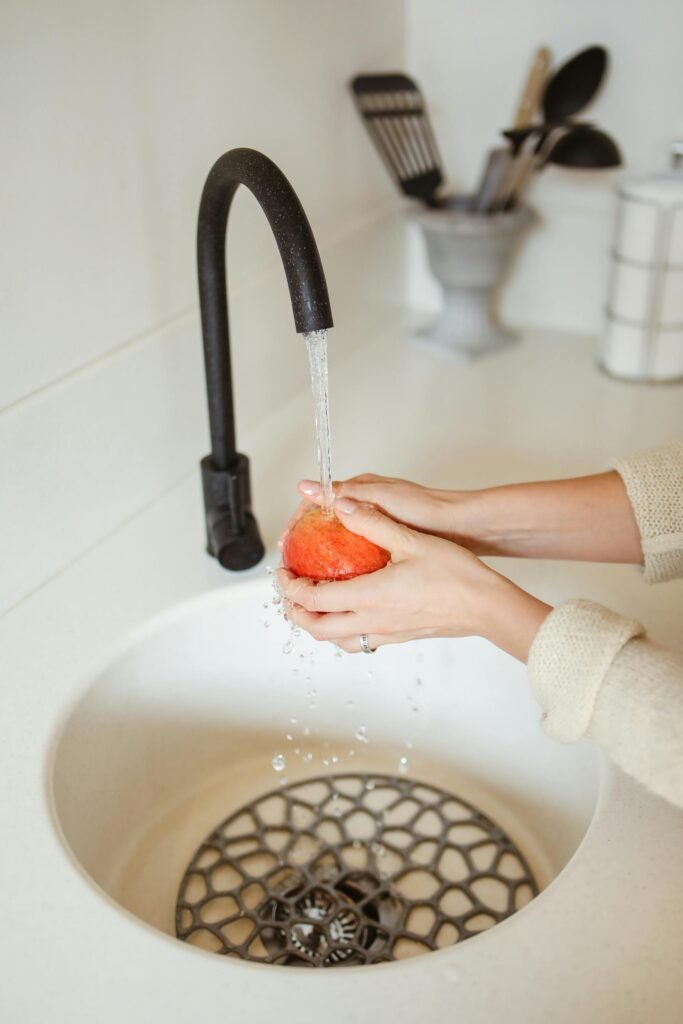 Close-up of hands washing a red apple under a black kitchen faucet.